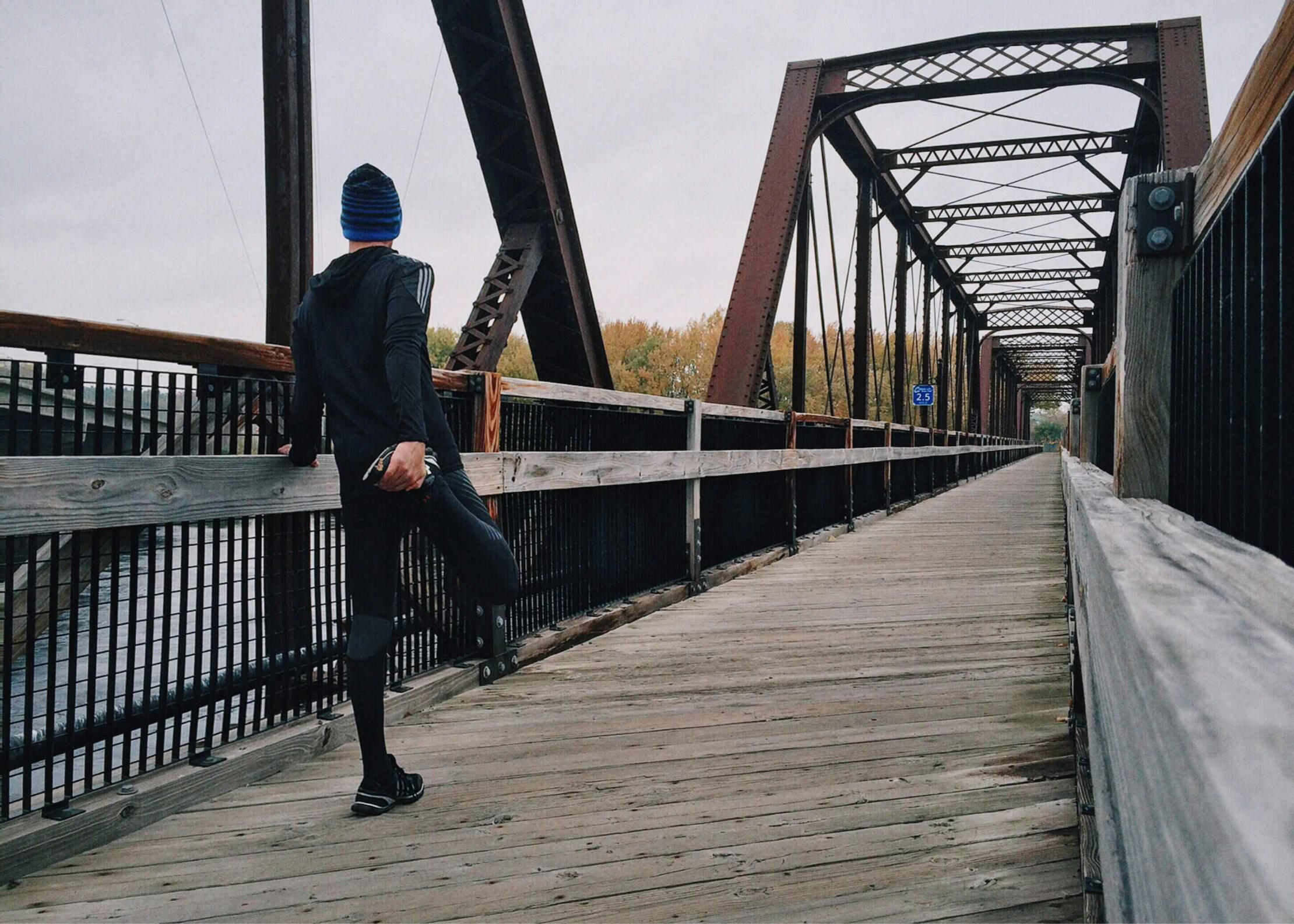 Athletic runner stretching their leg on a bridge railing, preparing for a morning workout with a forest skyline in the background.