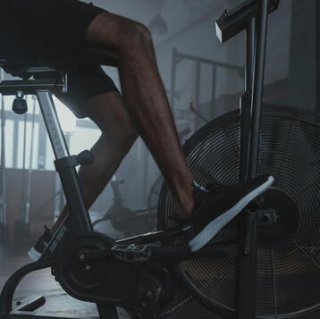 Man in athleisure riding a stationary bike, focused and pushing through a high-intensity workout in a dimly lit gym with ambient lighting.