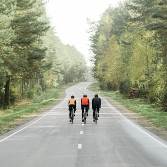 Three cyclists riding side by side on a road, with lush green forest flanking both sides, creating a scenic, natural backdrop.